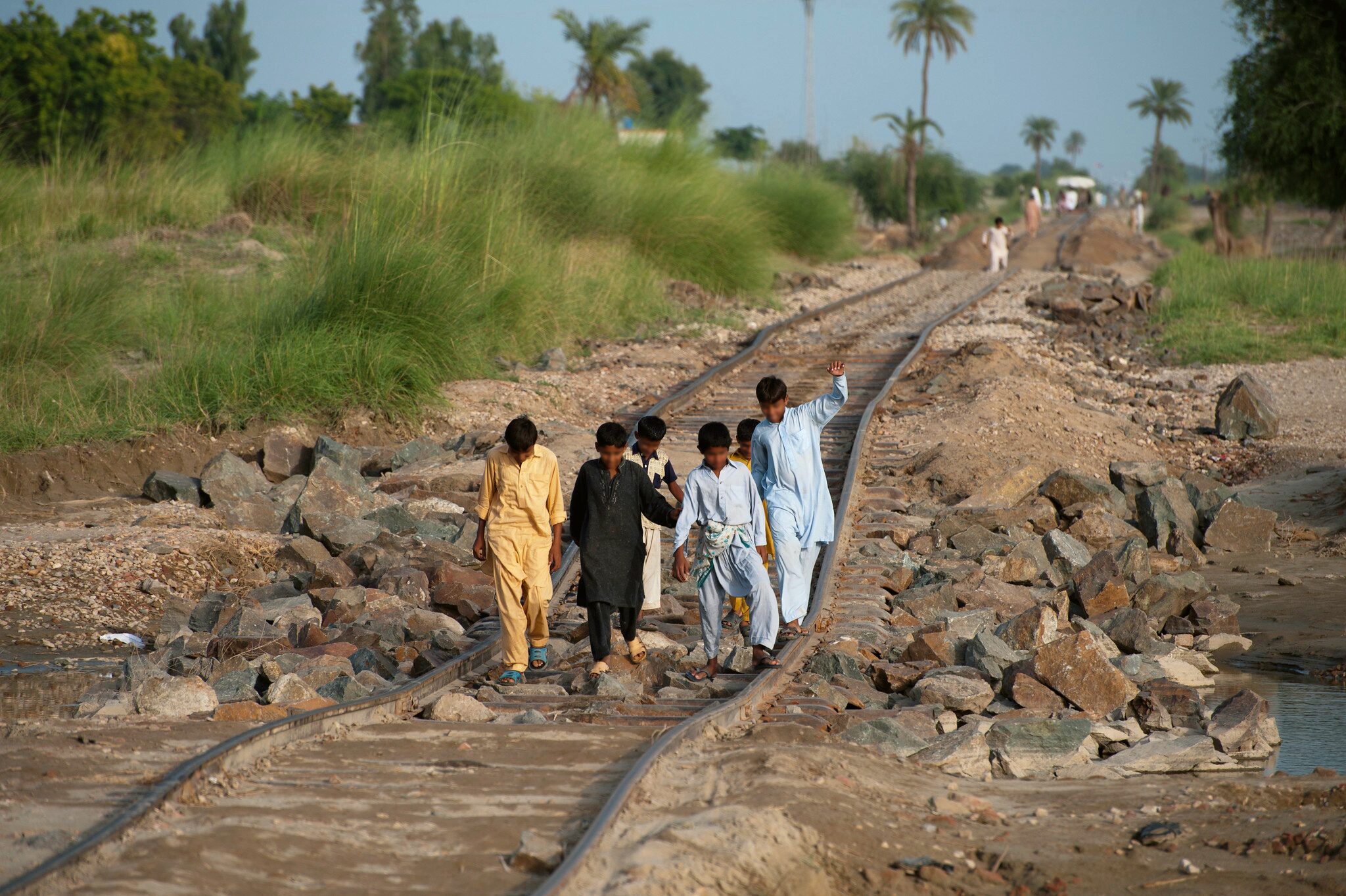 Image contains 6 young boys walking along train tracks in Pakistan. They are surrounded by plants and gravel, with water on the opposite side.